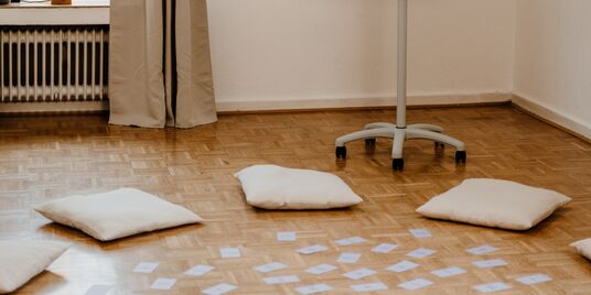 Heller Seminarraum. Auf dem Holzboden liegen mehrere Sitzkissen kreisförmig angeordnet, in der Mitte befinden sich Moderationskarten, Links ist ein Fenster mit Vorhang und Heizkörper zu sehen.